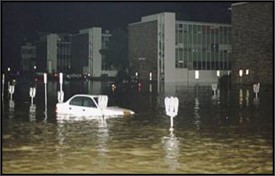 Photo showing flood waters in a parking lot, with a car submerged up to the hood.