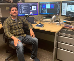 Candid photo of postdoctoral researcher Luis Aguilera seated at his desk with computer showing microscope images and programming code.