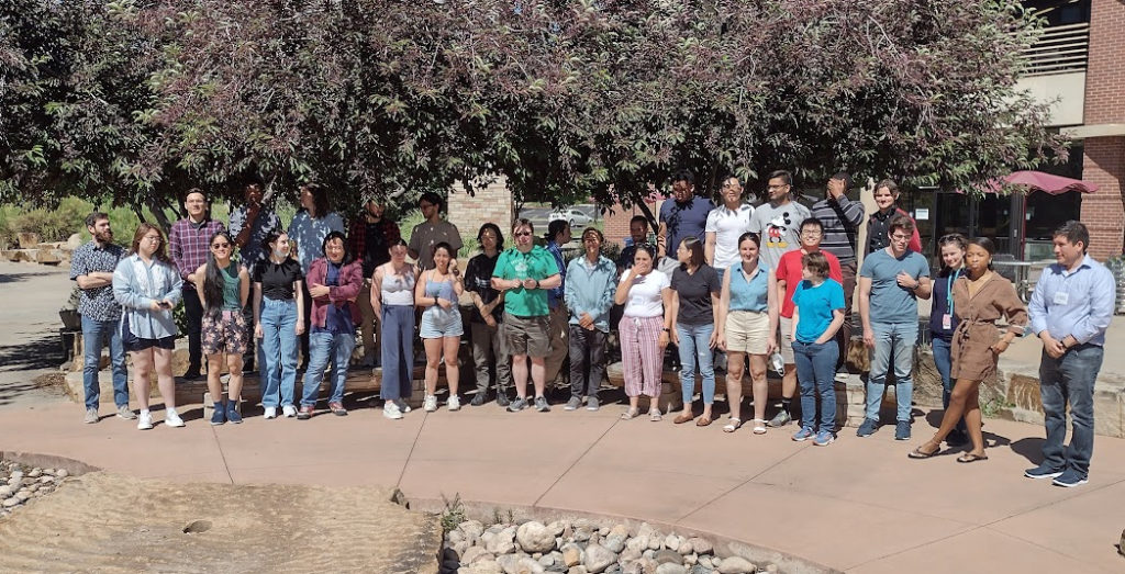 Outdoor group photo of UQ-Bio participants shows a couple dozen people in two rows beneath a flowering tree on the CSU campus.