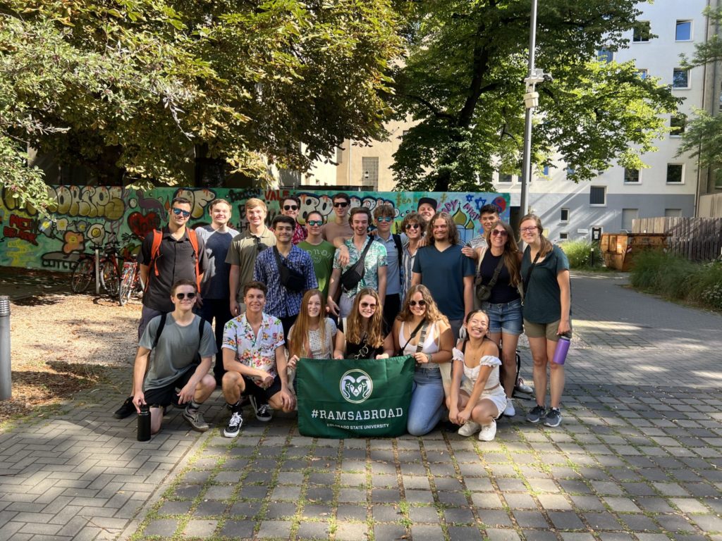 Engineering students studying abroad pose for a group photo outside their Berlin classroom with a flag bearing the CSU Ram logo and the text "#RAMSABROAD"