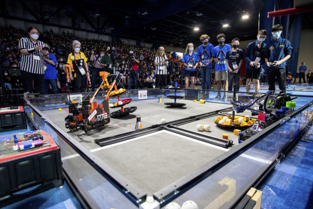 Robots compete in the foreground while referees and students controlling the robots look on.