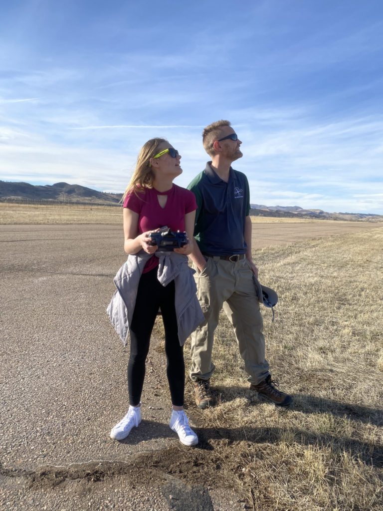 A young woman operates a drone as an instructor looks on. Background shows blue skies, foothills, and an empty airfield runway.