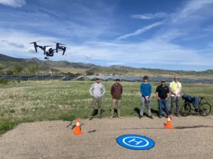 A drone hovers in front of a group of young men. Foothills and solar panels are visible in the background.