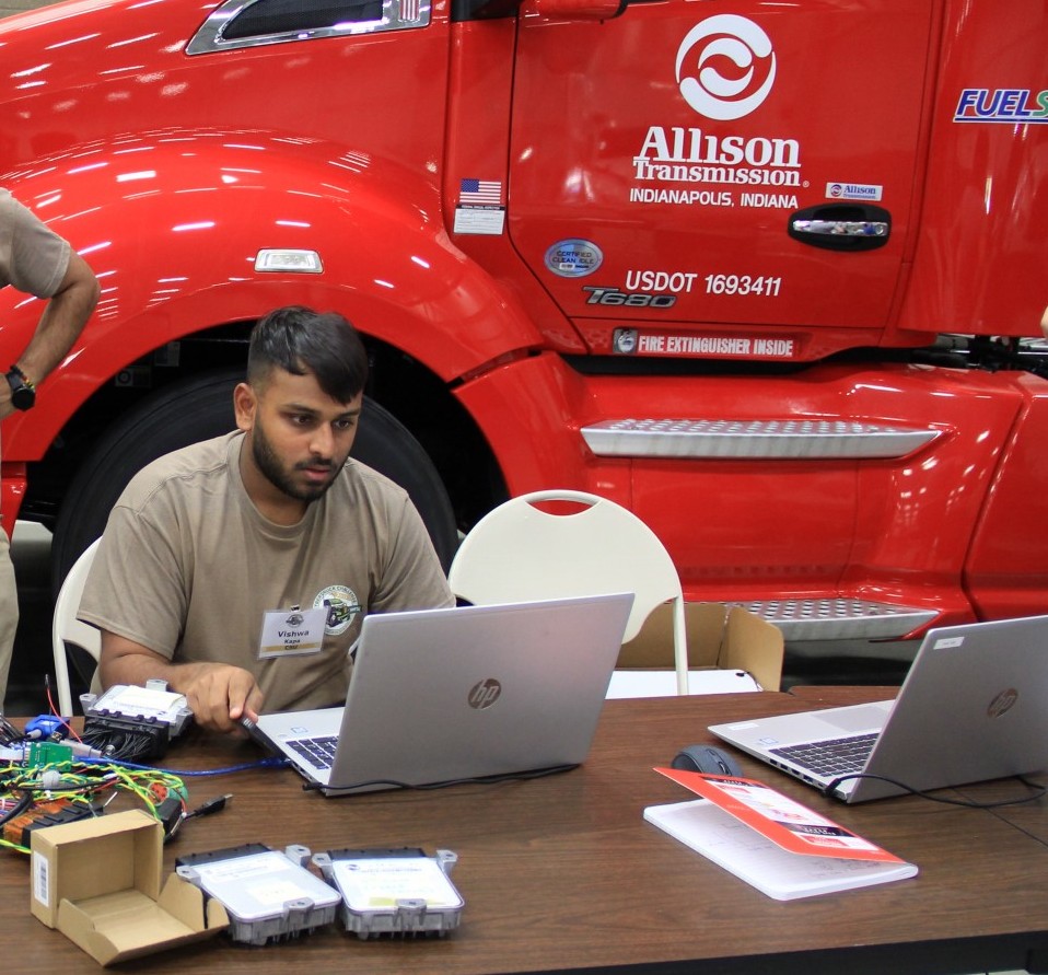 A student works on a laptop at a table with various electronic devices strewn about. In the background, a large red semi truck with a sign on the door reading "Allison Transmission".