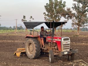 Photo of Kapa operating a red Massey Ferguson tractor in India. Foreground is tilled soil; vegetation and low foothills are visible in the haze in the background. 