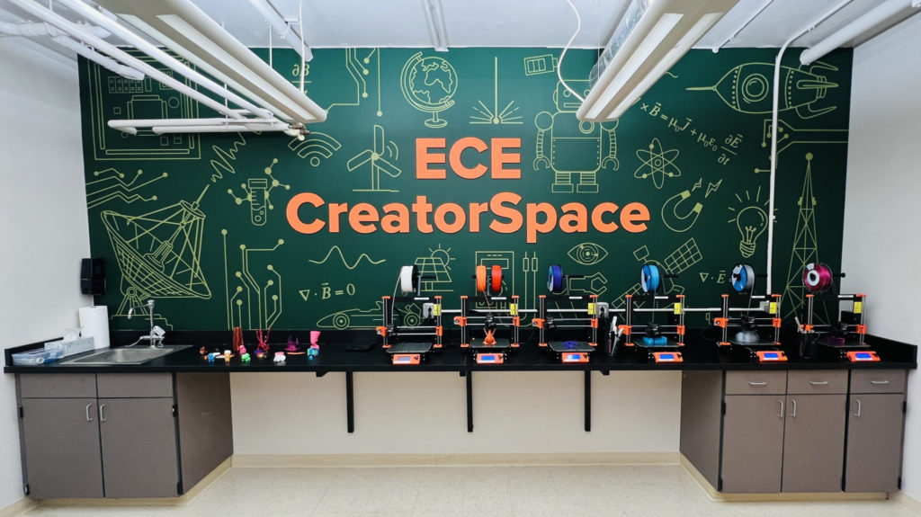 In the background, a prominent wall sign reading "ECE CreatorSpace" set in a graphic mimicking chalkboard drawings of radar dishes, robots, rockets, antennae, circuit boards, and alternative energy doodles. In the foreground, an array of 3D-printers and printed objects on a counter with a lab sink and cabinetry below.