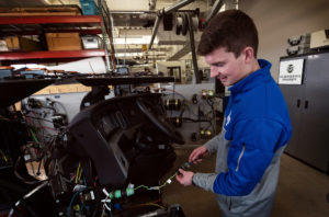 U.S. Air Force 2nd Lt. Gabe Salinger, a young white man with brown hair, holds up a wire connected to the internal components of a semi truck's cap that was removed and placed in a garage for testing. Behind him is the Colorado State University logo. He has on an Air Force Academy branded sweater.