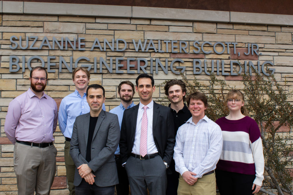 Group portrait of Yourdkhani with his research group, standing in front of a sandstone building with lettering reading "Suzanne and Walter Scott, Jr. Bioengineering Building"