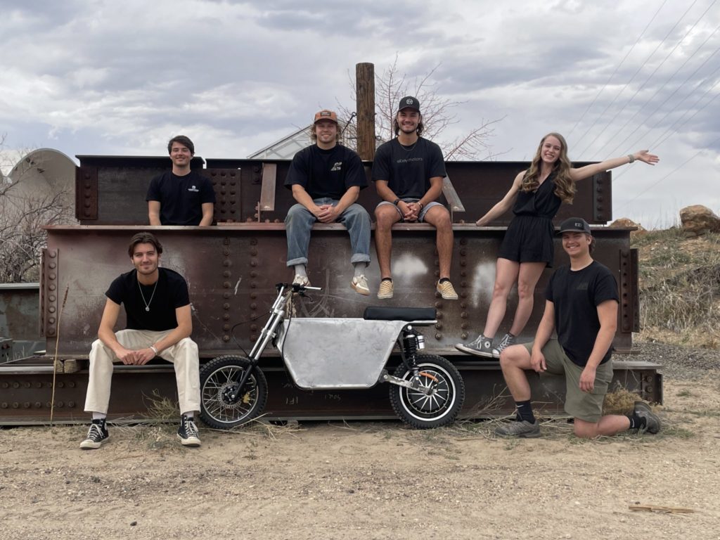 Students pose with a prototype electric motorcycle in front of a stack of large steel I-beams.