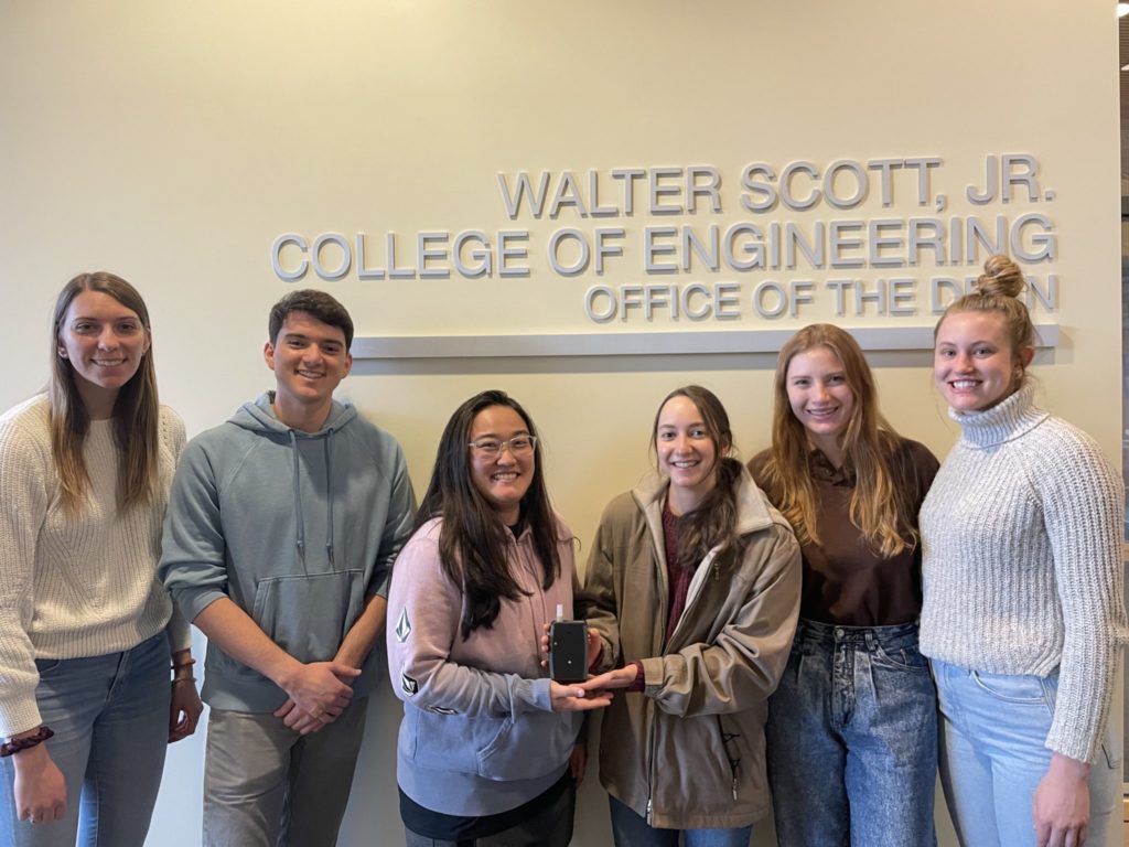 Members of the Sniftek team pose for an informal portrait in front of a wall sign reading "Walter Scott, Jr. College of Engineering" and "Office of the Dean"
