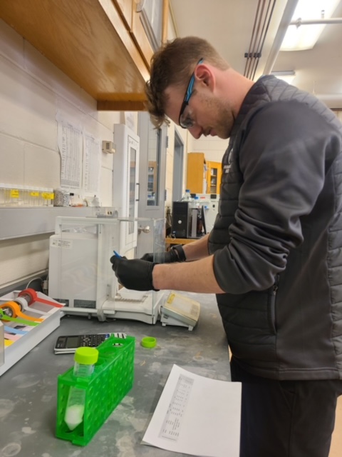 Wilson at work in a laboratory setting. He's wearing nitrile gloves and working with lab apparatus with a test tube rack and printed spreadsheet on the table nearby.