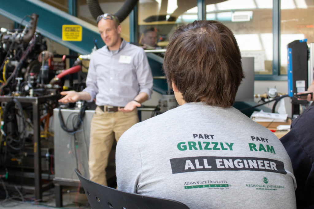 An instructor speaks in front of various engineering equipment. In the foreground, a student faces away from the camera, wearing a T-shirt that bears the text, "Part GRIZZLY/ Part RAM/ ALL ENGINEER." Below on the shirt are the CSU Mechanical Engineering department and Adams State University logos.