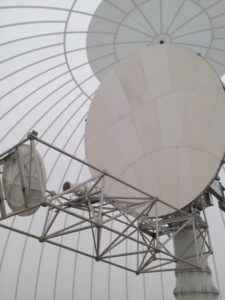 A photograph of a CSU-CHILL radar taken from inside the radome. In the foreground, a truss supports the radar emitter/receiver and a small reflector. A larger reflector sits behind the trust, mounted on the central pedestal. In the background, the curved ribs and skin of the radome are visible.