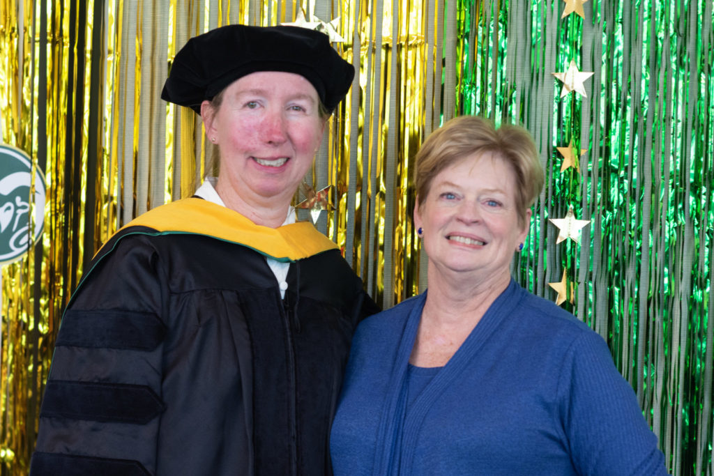 Sandra “Sandy” Dawson has on her doctoral regalia and is standing next to Ann Batchelor. Both are white women smiling. The background is strands of shiny party strings and stars in CSU colors. 