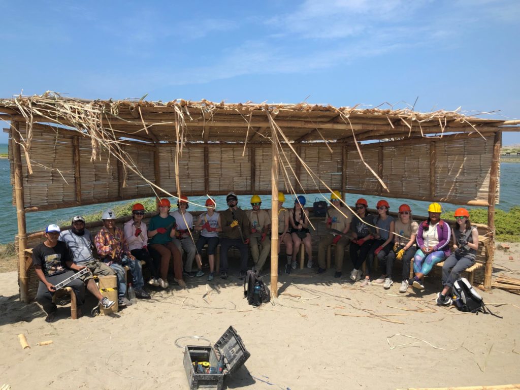 The group sits together in the shade of the completed bird blind, a rustic structure built from local bamboo and cane.