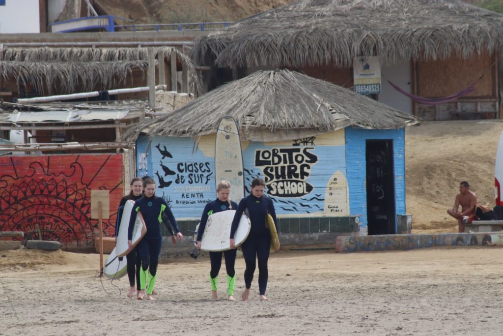 Students in wet suits carry surf boards down the beach. Behind them, a small thatched cinder block building bears a sign reading "Lobitos Surf School."