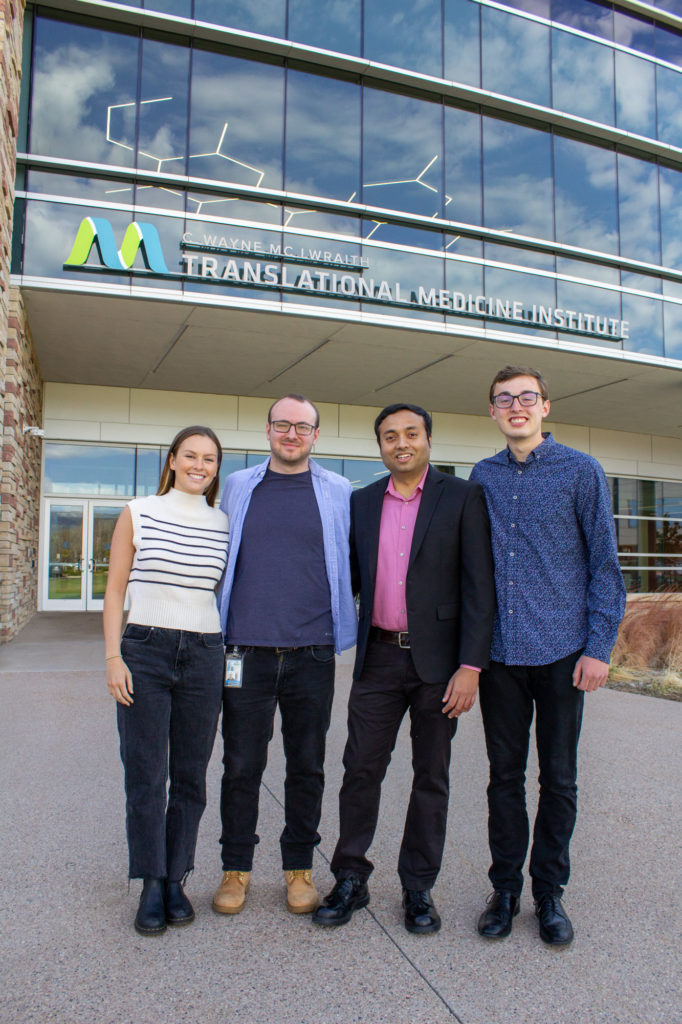 Four researchers stand in front of a large glass and sandstone building bearing a sign with a stylized DNA logo and the words "C Wayne McIlwraith Translational Medicine Institute". Visible through the glass is a light fixture in the form of an organic molecule diagram.