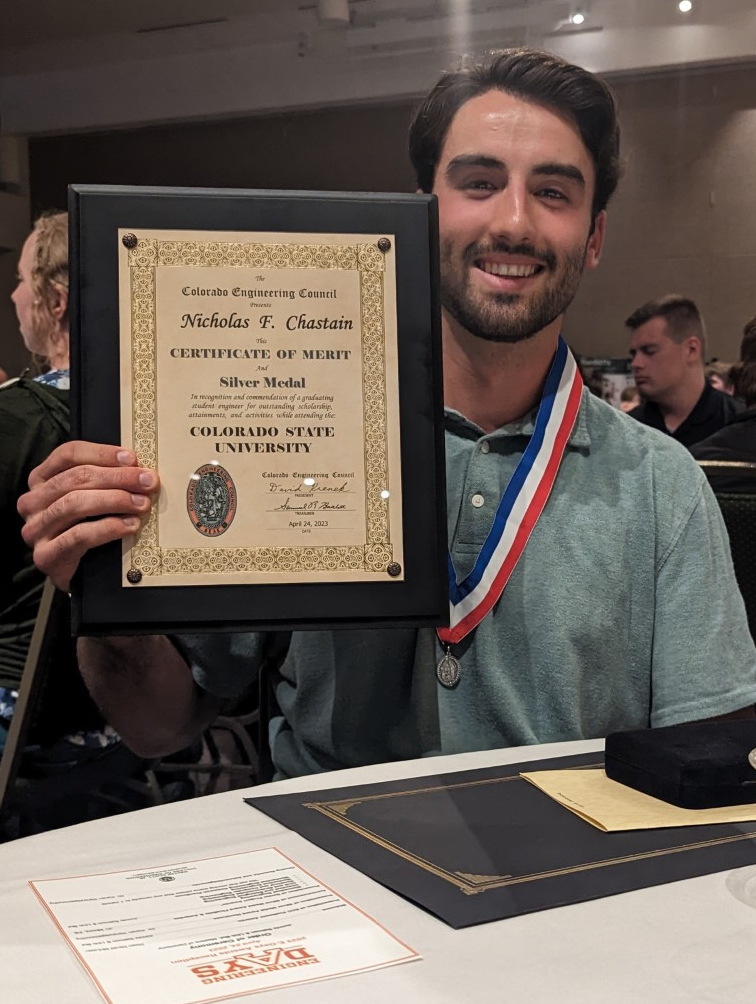 Nick Chastain holds a plaque reading, in part, "The Colorado Engineering Council presents Nicholas F. Chastain this Certificate of Merit and Silver Medal in recognition of...." Chastain also wears a red, white and blue ribbon bearing the silver medal around his neck. On the table is a program showing the CSU E-Days logo.