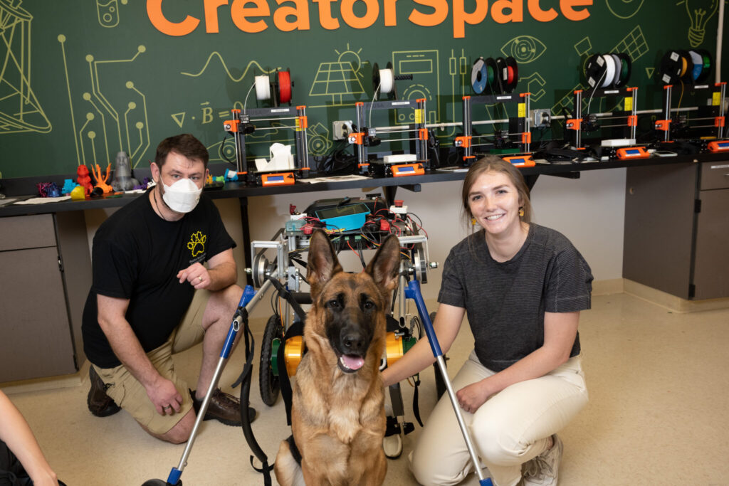 Two students pose for a casual portrait with a German shepherd in the exoskeleton apparatus