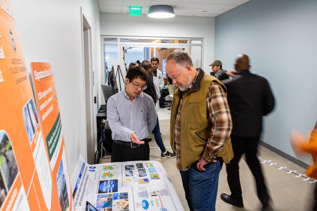 A candid photo from the CSU SPUR grand opening, in which Chen gestures at a table bearing posters and literature describing his research, while a member of the public listens and looks at the literature. 