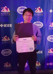 Haonan Chen poses for an informal portrait holding his award certificate, which bears the IEEE logo visible at the top. Behind him, a backdrop bears the logos of the IEEE, IEEE GRSS (Geoscience and Remote Sensing Society), and IGARSS Pasadena 2023.