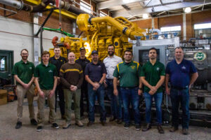 An informal indoor group portrait of 10 people from CSU, Caterpillar and Western Midstream. Behind the group sits a very large, yellow Caterpillar natural gas engine.