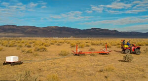 An outdoor photo showing an orange tractor towing a tTEM apparatus behind it. The landscape appears arid, with sparse, yellow and tan vegetation and brown foothills beneath a blue sky in the distance.
