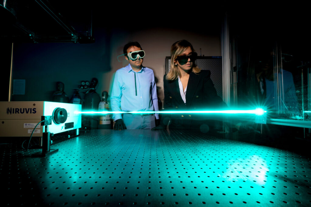 A male and female researcher in laser safety goggles look on as a laser flashes across the geometrically dotted space in front of them.