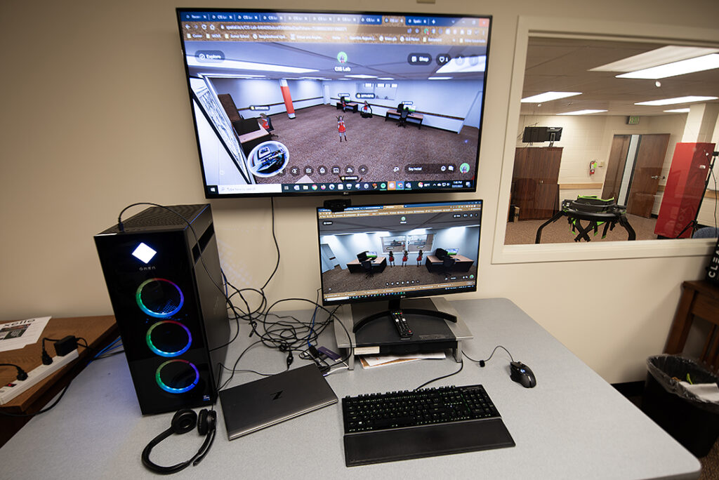The image is of a workspace with computer equipment. To the left, a window looks out to another room with a VR treadmill and padded support columns.