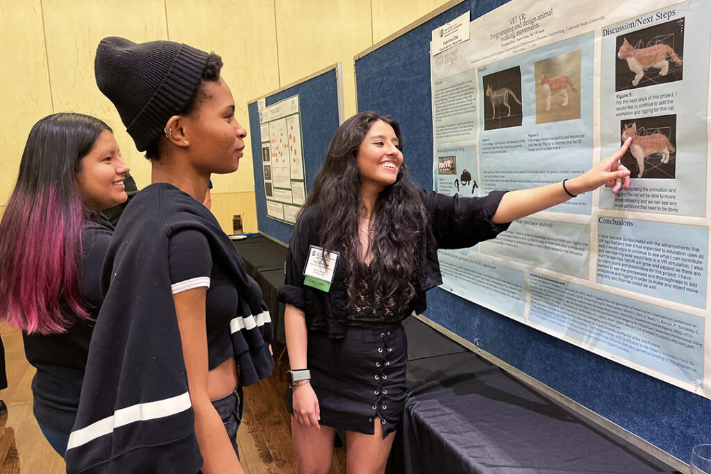 Valentina Diaz points at her poster with images of a simulated cat as two other students look on. The poster is titled: “VET VR: Programming and design animal walking movements.”