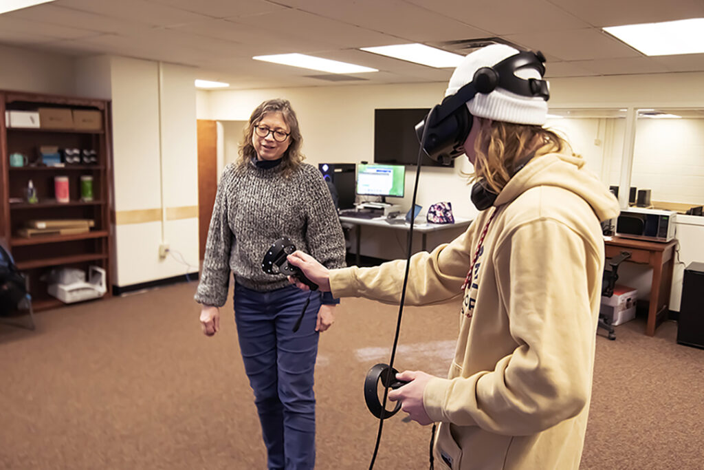 Marie Vans watches Evan Corwin as he uses virtual reality equipment including goggles and hand controls.