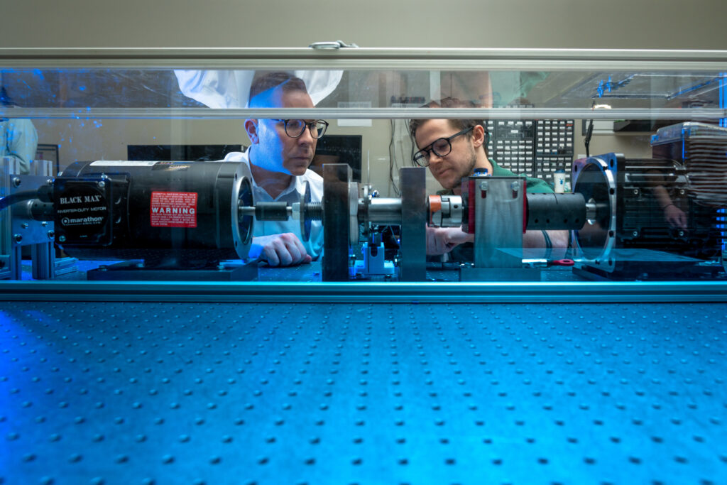 Two male researchers inspect test apparatus consisting of a large electric motor coupled to various other devices, all bolted to a perforated metallic laboratory table.