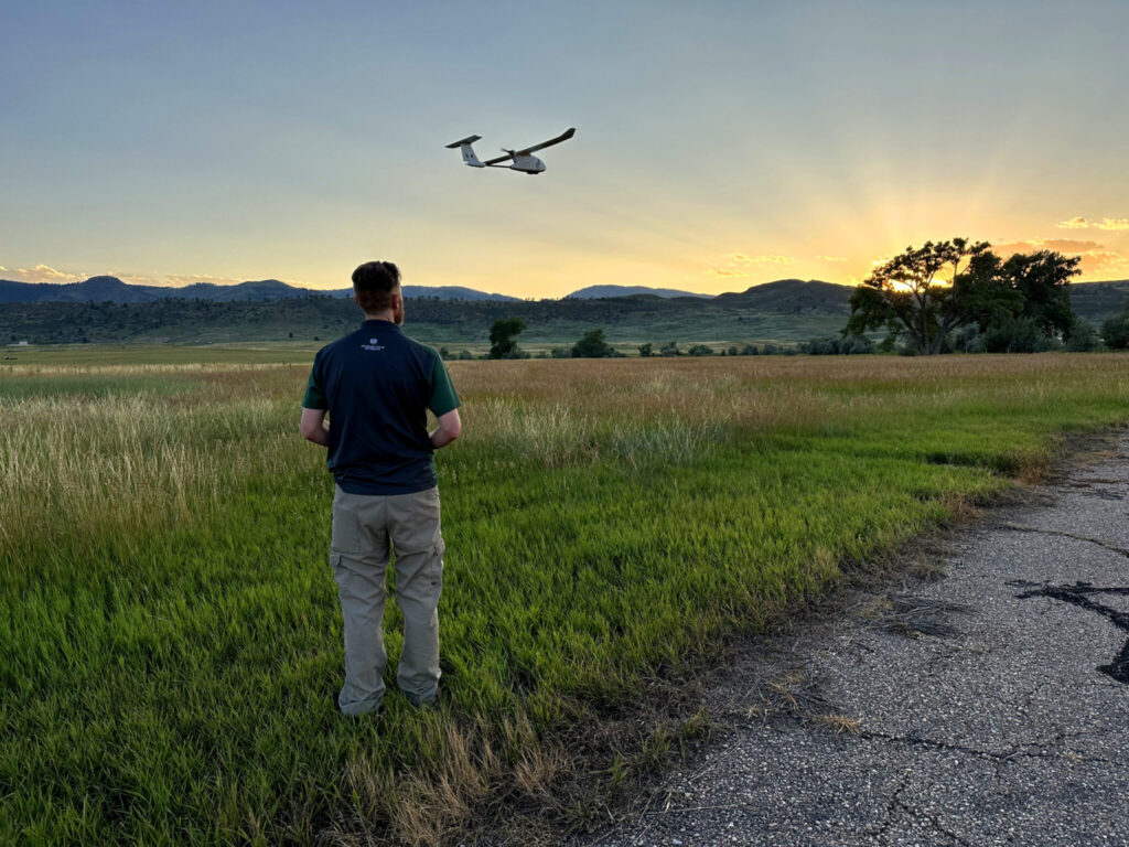 A man facing away from the camera stands on the grassy edge of a runway, operating a fixed-wing drone. In the background, the Front Range foothills and a large cottonwood tree are silhouetted against the sunset, and crepuscular rays reach skyward from the sun's location behind the mountains.