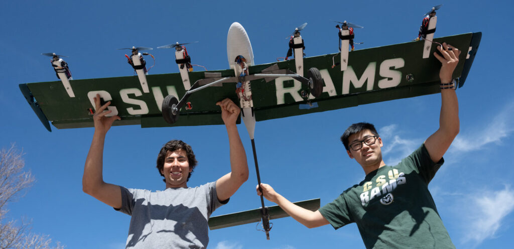 Two students hold a large fixed-wing drone that they built for a competition. The plane has 6 engines and a long tail boom. The wings are painted CSU green and bear the words "CSU RAMS" in white.