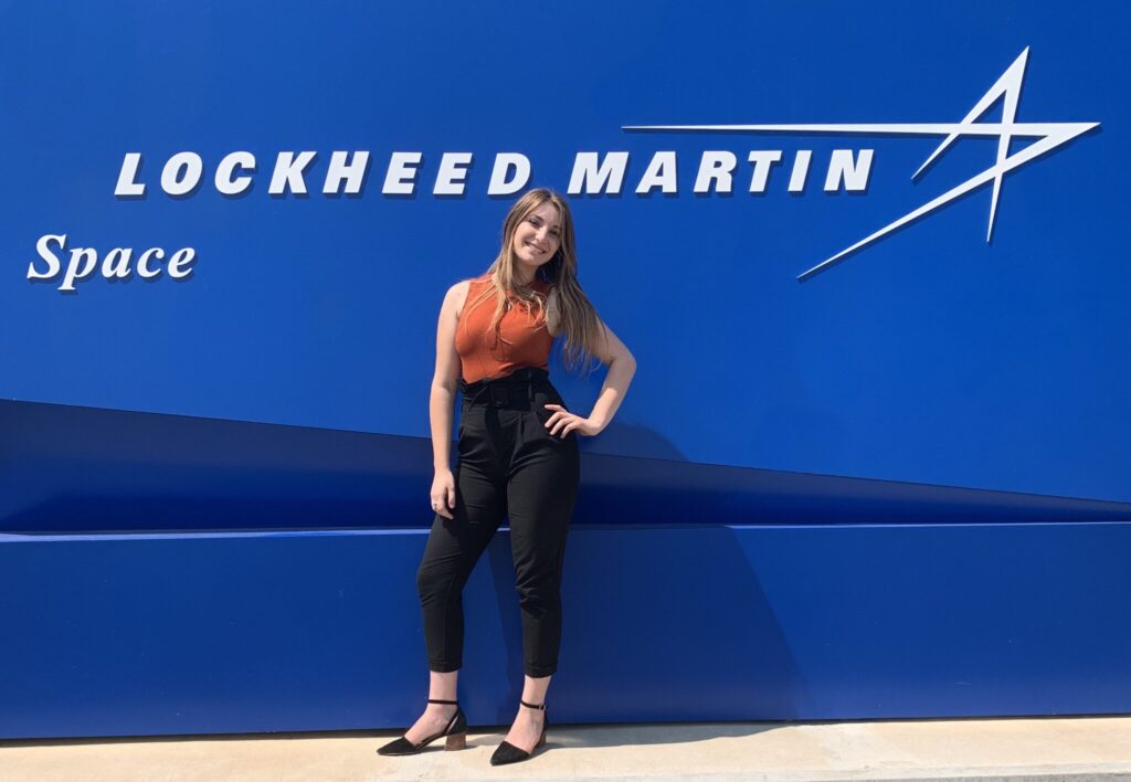 Kori Eliaz poses for a casual portrait outdoors in front of a large blue wall with the "Lockheed Martin Space" star logo on it.