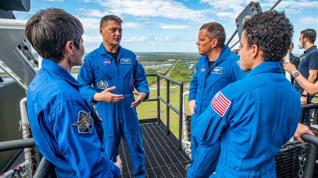 Candid photo showing 4 astronauts in blue flight suits with patches and insignia attached, standing on high tower at a launch facility. Dr. Kjell Lindgren gestures as he explains something to his colleagues.