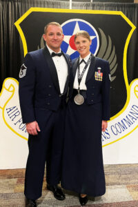 Mastrocola and her husband, a USAF Master Sergeant, pose together in dress uniform. Mastrocola wears a large silver medal hanging from a ribbon marked "AFSOC" and has a number of medals and service ribbons on her uniform. In the background, a partially obscured shield of the Air Force Special Operations Command is visible.