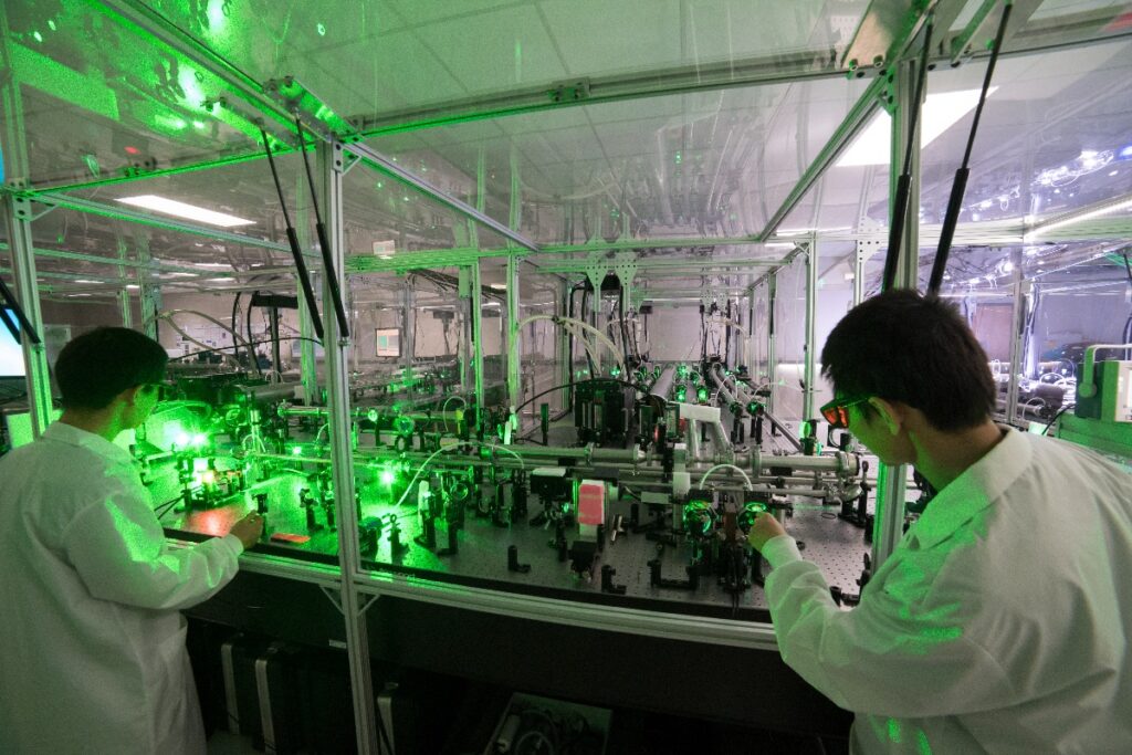 Researchers in lab coats work on a benchtop laser as its green light tints the laboratory.