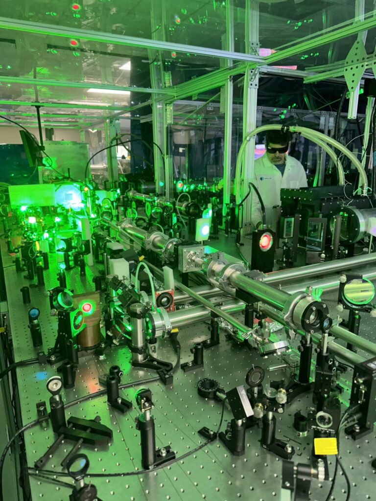 Laser apparatus on a lab table in the foreground, a white-coated researcher in laser safety goggles works with equipment while a green glow emanates from a target 