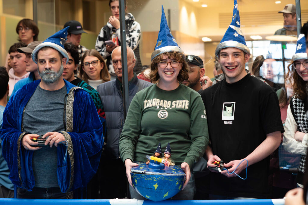 Three students dressed as wizards stand in front of a crowd, one student is holding a model-size blue boat.