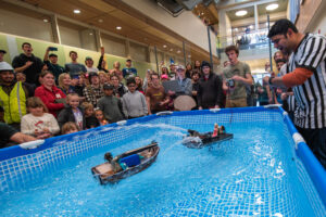 A crowd of people cheer on and record a miniature boat battle in a raised indoor pool. One of the boats sprays water at the other.