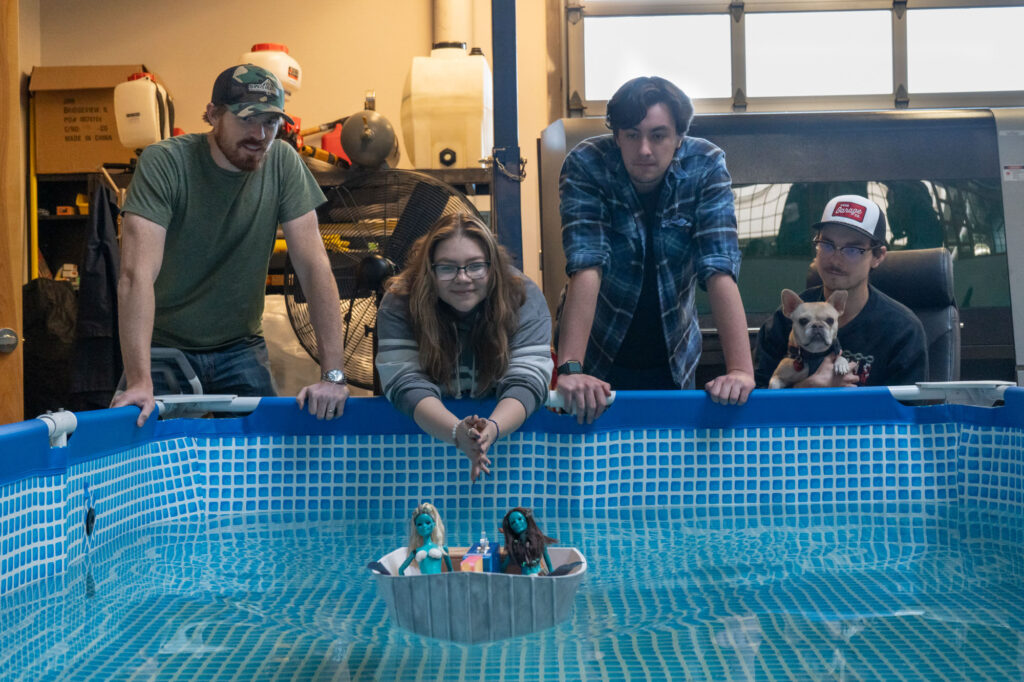Four students and a dog look over the edge of a raised indoor pool at a model boat floating on the water. The boat has two dolls spray painted blue to look like sirens.