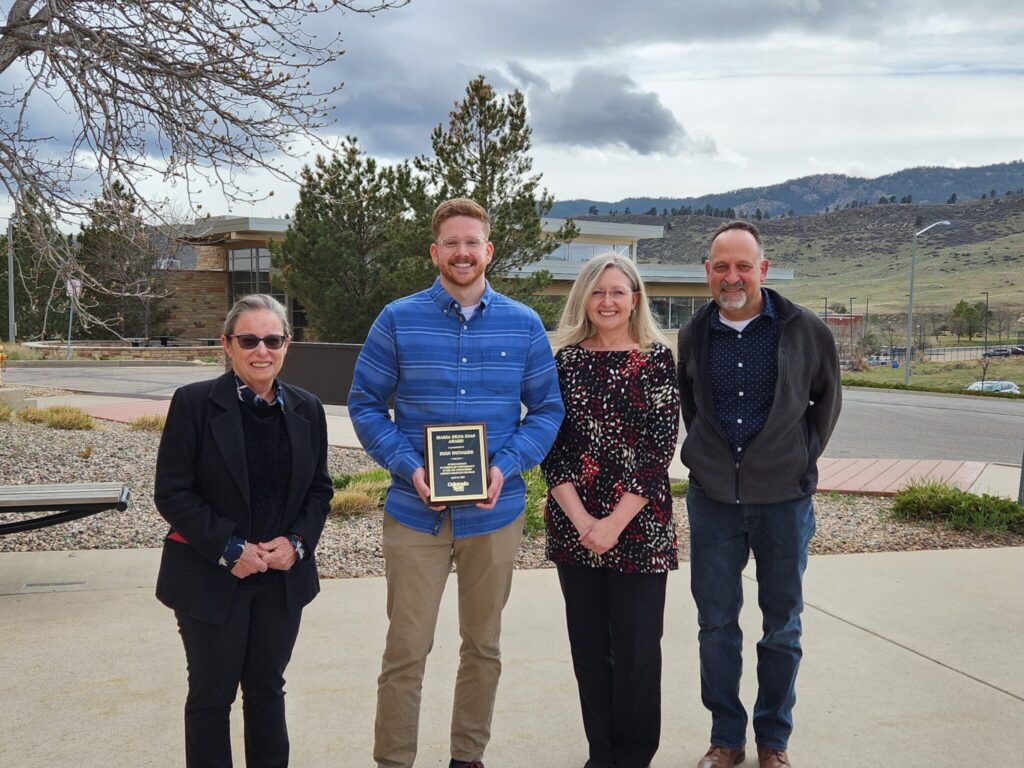 Two women and two men pose for a casual portrait outdoors near the Atmospheric Science building on the CSU Foothills Campus. The young man second from left holds a plaque in his hands.
