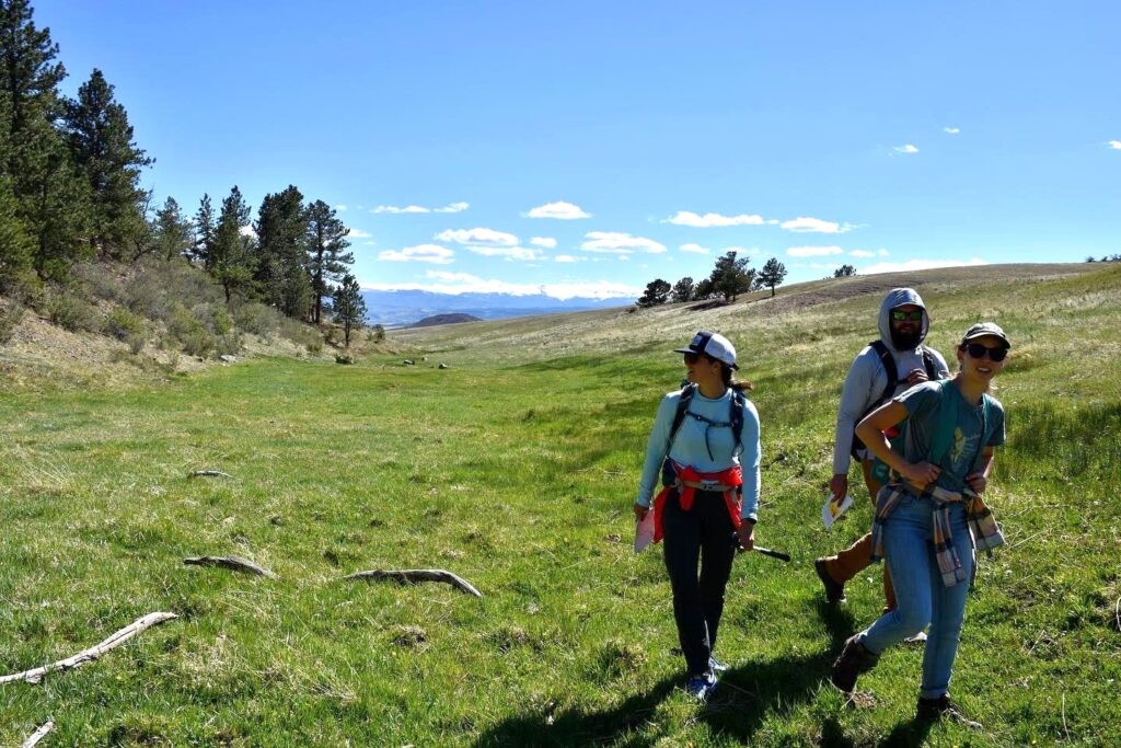 Three people with backpacks and hats walk in a green alpine meadow. The profile of the Rockies is visible in the distance beneath a blue sky.