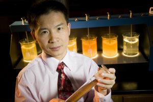 A researcher in shirt & tie holds a graduated cylinder with a murky liquid in it. In the background, several beakers sit on a table with lights suspended in their variously shaded liquids.