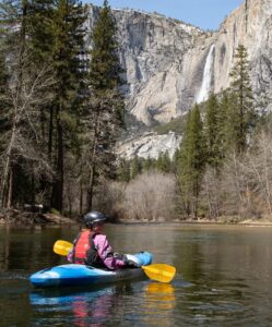 Davenport looks back over her right shoulder at the camera while sitting in a kayak on calm water. In the background, a tall, thin waterfall spills over a cliff face.