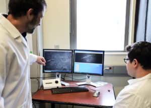 Two researchers in lb coats study a microscopic image of a single sperm cell on a computer monitor.