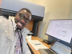 Oluwatobi “Tobi” Oke taking a smiling selfie seated at a desk in front of a laptop and open notebook 