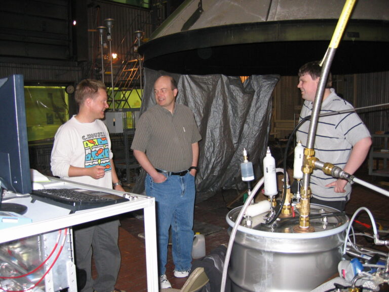 Collett and two other men talk in a laboratory setting. A large fume hood and a container with tubing and valve apparatus fill the foreground.