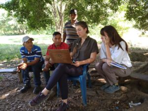 Student seated in chair outside under shade tree shows her laptop screen to individuals around her.